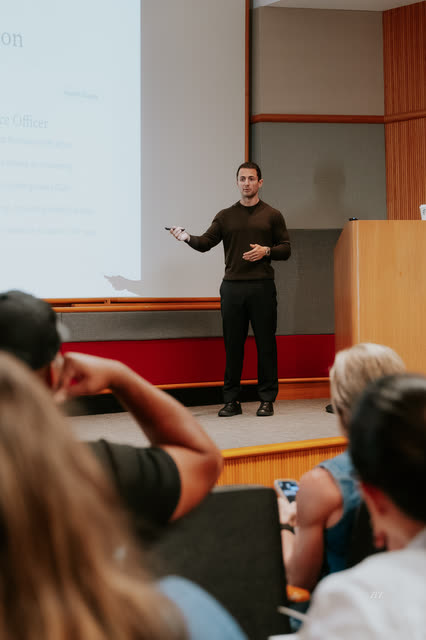 Speaker gesturing to the room, seen over attendees' shoulders