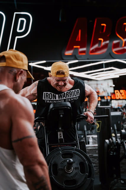 Coach spotting a T-bar row as the lifter in a COACH cap grinds out a rep
