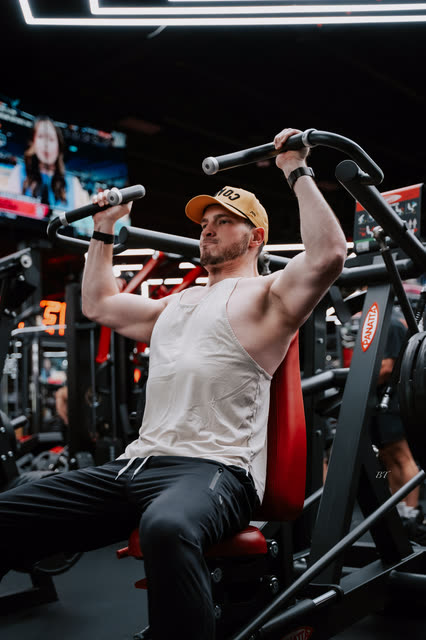Coach in a white tank and COACH cap pressing overhead on the shoulder press machine