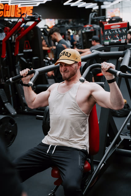 Coach in a white tank and COACH cap straining through a shoulder press