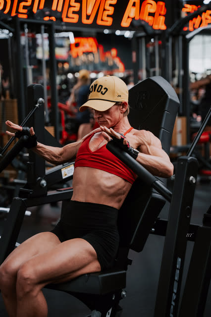 Coach in a red sports bra and COACH cap working a chest press machine