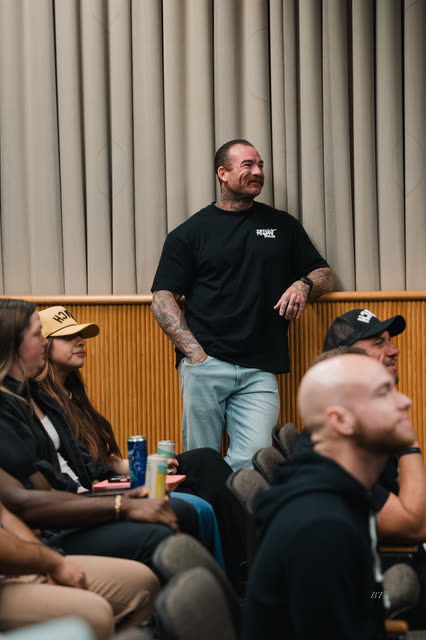 Coach with tattooed arms standing at the side of the lecture hall, smiling